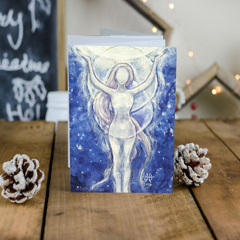 Moon Goddess Greeting Card and white envelope on rustic wooden table with pinecones  and lights behind